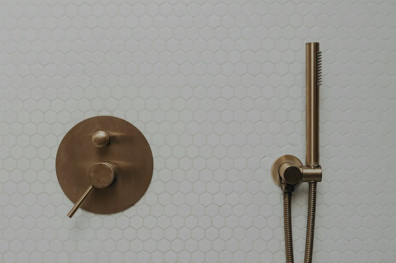 A brushed-brass shower valve and hand shower on white hex tile.