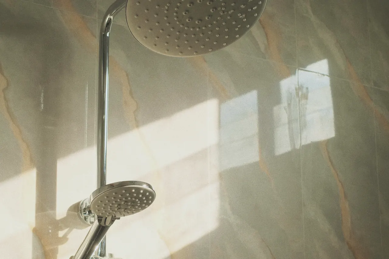 A chrome hand shower on a veined stone wall, lit by raking window sunlight.