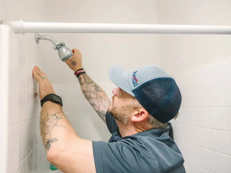 A technician in a company cap fits a new shower head on a white tiled wall.