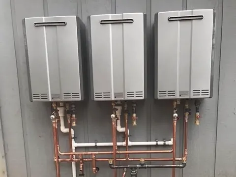 Three tankless water heaters mounted side by side on an outside wall, with new copper and PVC lines below.