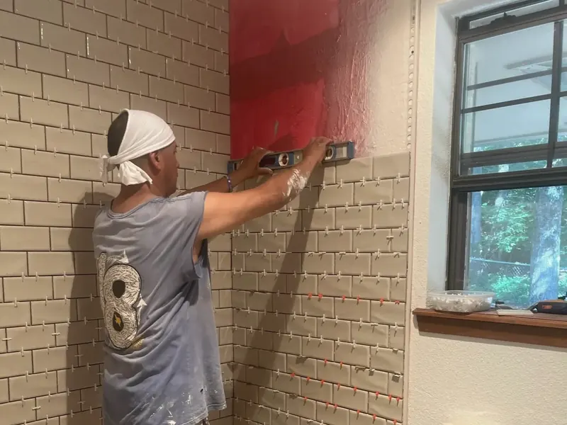 A worker sets tile on a bathroom wall above a tub, with tools and a bucket on the floor.