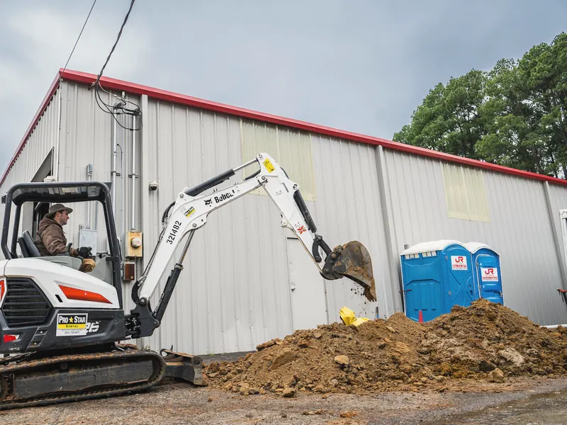 A mini excavator digs beside a metal building, with a pile of red dirt in front.