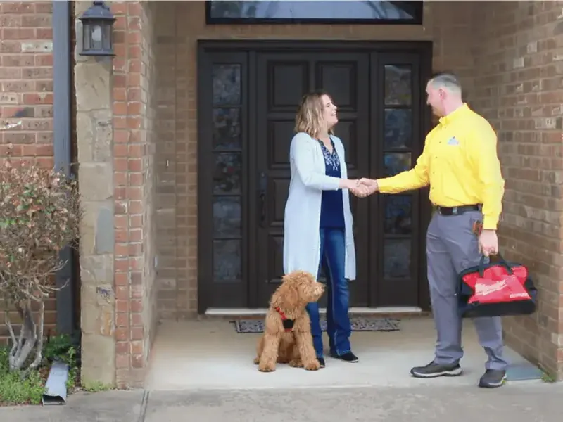 A technician in a yellow company shirt shakes hands with a homeowner at her front door; a dog sits beside them.