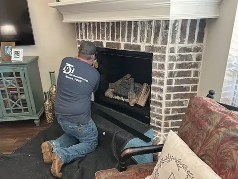 A technician in a company T-shirt kneels to service a gas log fireplace in a living room.